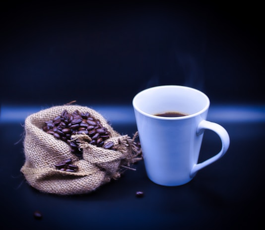 A steaming cup of coffee sits next to a burlap sack spilling over with roasted coffee beans, set against a dark background.