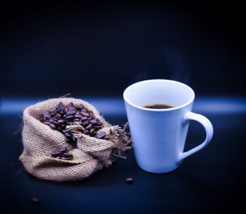 A steaming cup of coffee sits next to a burlap sack spilling over with roasted coffee beans, set against a dark background.