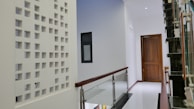 A modern indoor hallway with a glossy white floor and a minimalist design. The left side features a decorative white perforated wall, and there is a dark wooden door at the end of the hallway. A staircase with a glass and metal railing is visible on the right side.