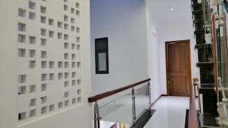 A modern indoor hallway with a glossy white floor and a minimalist design. The left side features a decorative white perforated wall, and there is a dark wooden door at the end of the hallway. A staircase with a glass and metal railing is visible on the right side.