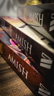 A close-up view of three books stacked on a wooden surface. Each book has 'AMISH' written on the spine in bold letters, with colorful covers and a glossy finish. The books are slightly tilted, creating a dynamic composition with shadows and light reflections.
