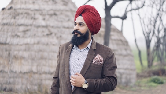 A man with a beard wearing a red turban and a brown suit stands outdoors. He has a striped shirt underneath and a pocket square in his breast pocket. Behind him, there are blurred images of what appear to be thatched huts and bare trees, suggesting a rural setting.