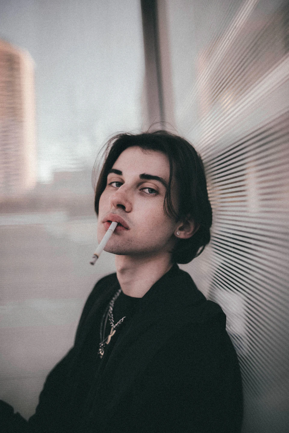 Young man sits against a wall, holding a cigarette, with dark clothing and long hair