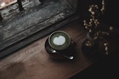 A cup of matcha latte with a heart-shaped latte art on top, placed on a wooden surface beside a window with a rustic and weathered look. A small vase with dried flowers is also placed on the same surface, and a spoon rests on the saucer under the cup.