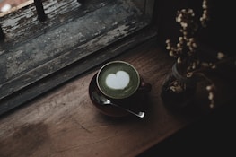 A cup of matcha latte with a heart-shaped latte art on top, placed on a wooden surface beside a window with a rustic and weathered look. A small vase with dried flowers is also placed on the same surface, and a spoon rests on the saucer under the cup.