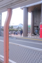 A person in business attire is walking toward a building with tall pillars. There are bicycles parked in a bike rack area, and a red sign with text near the entrance. A road with a white metal fence is in the foreground, and buildings are visible in the background.