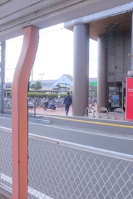 A person in business attire is walking toward a building with tall pillars. There are bicycles parked in a bike rack area, and a red sign with text near the entrance. A road with a white metal fence is in the foreground, and buildings are visible in the background.