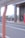 A person in business attire is walking toward a building with tall pillars. There are bicycles parked in a bike rack area, and a red sign with text near the entrance. A road with a white metal fence is in the foreground, and buildings are visible in the background.