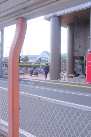 A person in business attire is walking toward a building with tall pillars. There are bicycles parked in a bike rack area, and a red sign with text near the entrance. A road with a white metal fence is in the foreground, and buildings are visible in the background.
