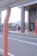 A person in business attire is walking toward a building with tall pillars. There are bicycles parked in a bike rack area, and a red sign with text near the entrance. A road with a white metal fence is in the foreground, and buildings are visible in the background.