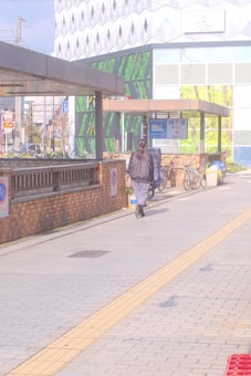 A person walks along a paved sidewalk with a yellow tactile guidance path. The surroundings include a modern building with geometric patterns and a mix of green and white panels. Bicycles are parked next to a low brick wall, and there are clear sunny skies above.