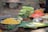An assortment of fresh fruits and vegetables arranged on a table. There are sliced cucumbers, bunches of green vegetables, diced papayas, and boiled sweet corn. Some lemons are also visible, along with cut orange sections.