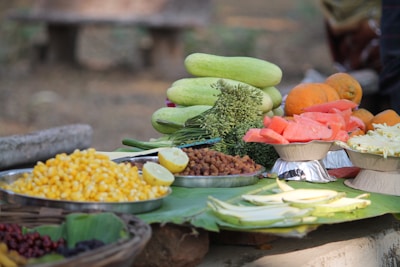 An assortment of fresh fruits and vegetables arranged on a table. There are sliced cucumbers, bunches of green vegetables, diced papayas, and boiled sweet corn. Some lemons are also visible, along with cut orange sections.