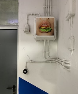 A wall with electrical conduits and a painted picture of lips holding a hamburger placed within a frame. The wall is primarily white with a section painted blue, and a gray door is visible beside the art piece. Several pipes and wires are running along the wall, some with visible connectors and bends.
