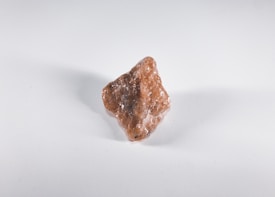 A rough, unpolished reddish-brown mineral or rock sits on a plain white background. The surface of the rock is uneven and appears to have some crystalline textures, with small flecks of white visible on its surface.