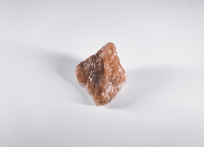 A rough, unpolished reddish-brown mineral or rock sits on a plain white background. The surface of the rock is uneven and appears to have some crystalline textures, with small flecks of white visible on its surface.