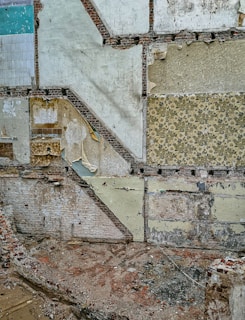 A partially demolished building wall with layers of exposed old wallpaper and bricks. The wall displays various patterns and textures, with some areas revealing worn-out paints and plaster. The ground is littered with rubble and debris.