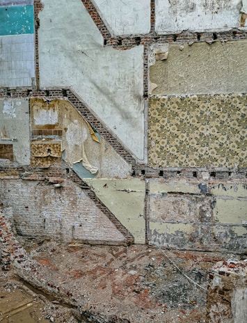 A partially demolished building wall with layers of exposed old wallpaper and bricks. The wall displays various patterns and textures, with some areas revealing worn-out paints and plaster. The ground is littered with rubble and debris.