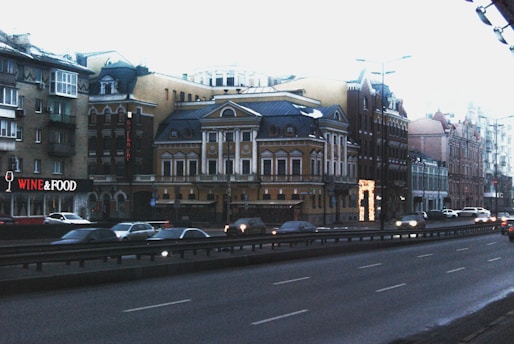 A city street bordered by a row of buildings with classic European architecture. The street is busy with moving cars and the sky above is overcast. One of the buildings features a sign that says 'WINE & FOOD'.