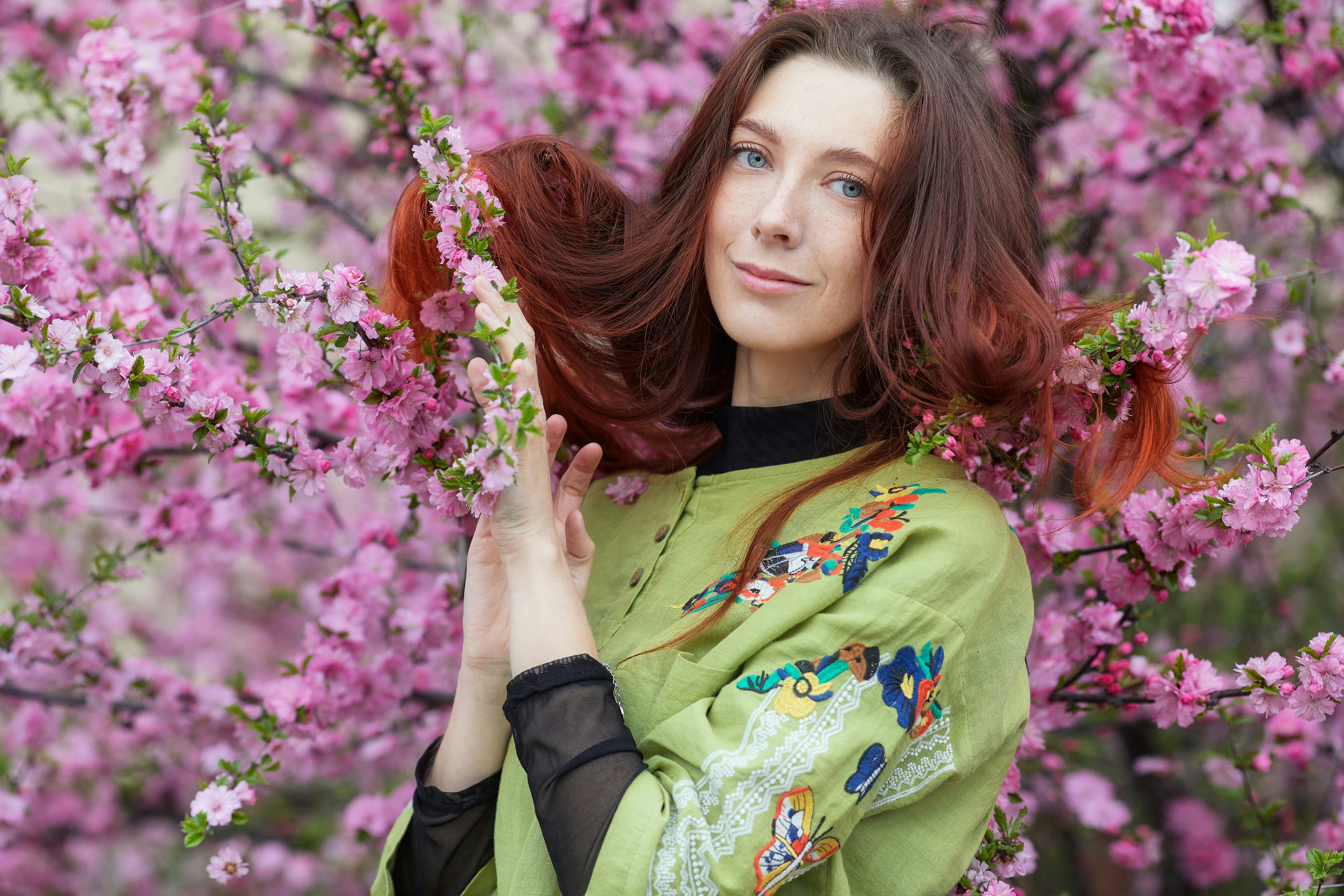 Portrait of a sensual red-haired young woman in a delicate spring garden. Beautiful girl in green clothes and long red hair posing against a background of gorgeous pink flowers