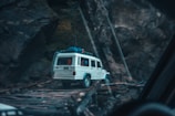 A white SUV is seen navigating a narrow, wooden bridge set in a rugged, rocky landscape. The vehicle carries luggage on its roof and is surrounded by tall, dark rock formations.