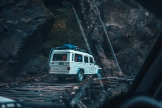 A white SUV is seen navigating a narrow, wooden bridge set in a rugged, rocky landscape. The vehicle carries luggage on its roof and is surrounded by tall, dark rock formations.