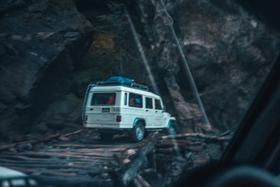 A white SUV is seen navigating a narrow, wooden bridge set in a rugged, rocky landscape. The vehicle carries luggage on its roof and is surrounded by tall, dark rock formations.