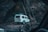 A white SUV is seen navigating a narrow, wooden bridge set in a rugged, rocky landscape. The vehicle carries luggage on its roof and is surrounded by tall, dark rock formations.