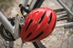 A close-up view of a bicycle featuring a red helmet attached to the frame. The bicycle is silver-gray, and its pedal and chain are visible. The background is slightly blurred, hinting at a gravel or stone surface.