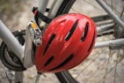 A close-up view of a bicycle featuring a red helmet attached to the frame. The bicycle is silver-gray, and its pedal and chain are visible. The background is slightly blurred, hinting at a gravel or stone surface.