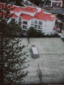 Aerial view of a residential area featuring white buildings with red-tiled roofs surrounded by greenery. In the foreground, a fenced tennis court with a small structure is visible, set amidst tall pine trees.