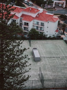Aerial view of a residential area featuring white buildings with red-tiled roofs surrounded by greenery. In the foreground, a fenced tennis court with a small structure is visible, set amidst tall pine trees.