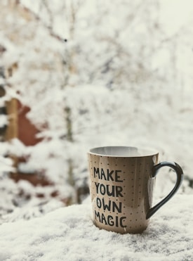 A ceramic mug with the phrase 'MAKE YOUR OWN MAGIC' printed on it is placed on a snow-covered surface. The background is filled with snow-covered trees and an out-of-focus building.