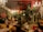 An ornate room is filled with lush greenery, intricate chandeliers, and an eclectic mix of decorative statues and vintage furniture. The space features a warm, ambient lighting with elegant chandeliers hanging from a partially exposed wooden ceiling. Several potted plants and hanging ornaments create an exotic and vibrant atmosphere.