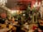 An ornate room is filled with lush greenery, intricate chandeliers, and an eclectic mix of decorative statues and vintage furniture. The space features a warm, ambient lighting with elegant chandeliers hanging from a partially exposed wooden ceiling. Several potted plants and hanging ornaments create an exotic and vibrant atmosphere.