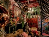 A richly decorated room features an eclectic mix of antique and vintage furniture and decor. A prominent painting of a woman in an ornate frame hangs on the wall surrounded by lush greenery and vibrant floral arrangements. A statue of a seated figure with a plumed hat sits on an intricately carved sofa, adorned with various patterned cushions. An elaborate chandelier with crystal details hangs from the ceiling, and a red lacquered cabinet with Asian motifs stands in the background. Various decorative elements, including hanging lights and plants, create a warm and inviting atmosphere.