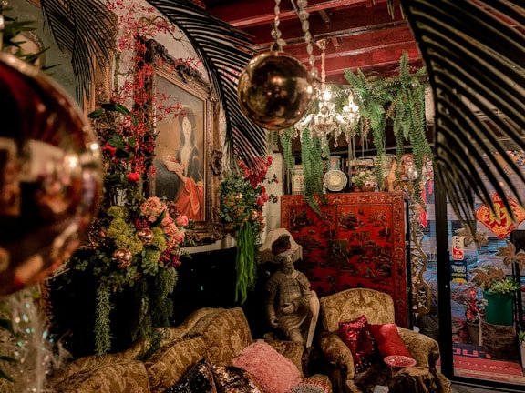 A richly decorated room features an eclectic mix of antique and vintage furniture and decor. A prominent painting of a woman in an ornate frame hangs on the wall surrounded by lush greenery and vibrant floral arrangements. A statue of a seated figure with a plumed hat sits on an intricately carved sofa, adorned with various patterned cushions. An elaborate chandelier with crystal details hangs from the ceiling, and a red lacquered cabinet with Asian motifs stands in the background. Various decorative elements, including hanging lights and plants, create a warm and inviting atmosphere.