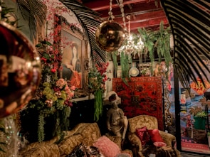 A richly decorated room features an eclectic mix of antique and vintage furniture and decor. A prominent painting of a woman in an ornate frame hangs on the wall surrounded by lush greenery and vibrant floral arrangements. A statue of a seated figure with a plumed hat sits on an intricately carved sofa, adorned with various patterned cushions. An elaborate chandelier with crystal details hangs from the ceiling, and a red lacquered cabinet with Asian motifs stands in the background. Various decorative elements, including hanging lights and plants, create a warm and inviting atmosphere.