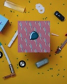 A vibrant composition featuring a box adorned with a pattern of small, colorful birds on pink, set against a yellow background. Scattered around are various objects, including a toy car, a plastic ring, a tube, and other small items, creating an eclectic and playful arrangement.