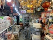An interior of a crowded shop filled with various household and electrical items, including light bulbs, cables, and packaged goods. Red lanterns with tassels hang from the ceiling, along with ornate chandeliers. Shelves and displays are lined with boxes and products, and the space is brightly lit.