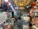 An interior of a crowded shop filled with various household and electrical items, including light bulbs, cables, and packaged goods. Red lanterns with tassels hang from the ceiling, along with ornate chandeliers. Shelves and displays are lined with boxes and products, and the space is brightly lit.
