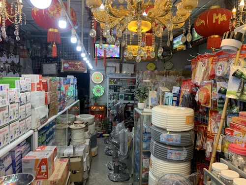 An interior of a crowded shop filled with various household and electrical items, including light bulbs, cables, and packaged goods. Red lanterns with tassels hang from the ceiling, along with ornate chandeliers. Shelves and displays are lined with boxes and products, and the space is brightly lit.
