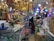 A store is filled with a variety of lighting fixtures such as chandeliers and lamps, which are displayed hanging from the ceiling. Numerous boxes and packages, likely containing electronic or lighting equipment, are neatly stacked on shelves and counters. Two people are visible in the image, surrounded by the merchandise. A potted plant and other miscellaneous items add to the cluttered yet organized feel of the space.