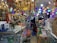 A store is filled with a variety of lighting fixtures such as chandeliers and lamps, which are displayed hanging from the ceiling. Numerous boxes and packages, likely containing electronic or lighting equipment, are neatly stacked on shelves and counters. Two people are visible in the image, surrounded by the merchandise. A potted plant and other miscellaneous items add to the cluttered yet organized feel of the space.