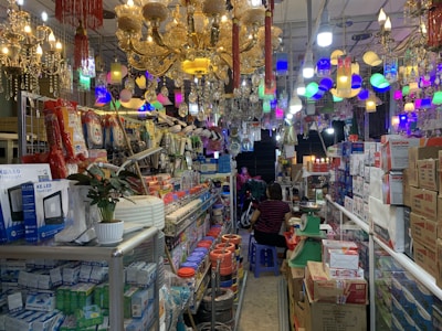 A store is filled with a variety of lighting fixtures such as chandeliers and lamps, which are displayed hanging from the ceiling. Numerous boxes and packages, likely containing electronic or lighting equipment, are neatly stacked on shelves and counters. Two people are visible in the image, surrounded by the merchandise. A potted plant and other miscellaneous items add to the cluttered yet organized feel of the space.