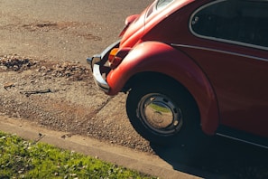 A classic red Volkswagen Beetle is parked by the side of a paved road, partially on the grass. The rear part of the car is visible, showcasing its distinctive rounded shape and chrome details. Sunlight casts shadows, highlighting the contours of the vehicle and creating a nostalgic ambiance.