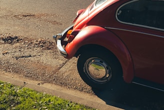 A classic red Volkswagen Beetle is parked by the side of a paved road, partially on the grass. The rear part of the car is visible, showcasing its distinctive rounded shape and chrome details. Sunlight casts shadows, highlighting the contours of the vehicle and creating a nostalgic ambiance.