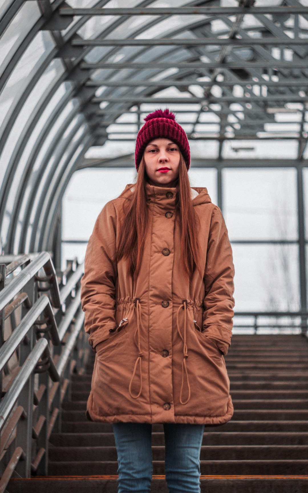woman in red knit cap and brown coat standing on gray metal bridge
