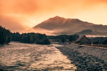 A serene landscape featuring a flowing river bordered by a rocky bank on one side and a row of wooden cabins on the other. In the background, a range of mountains looms under a golden sky tinged with hues of orange, creating a warm and tranquil atmosphere. Mist lingers over the distant trees, enhancing the peaceful ambiance.