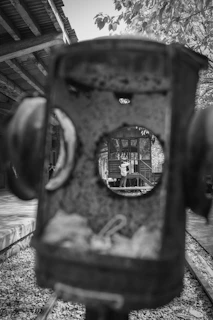 A black and white photograph taken through the circular opening of an old, rusted train signal. In the background, there is a person standing on a railway platform, focusing a camera, with an old wooden building and overhanging tree branches visible.